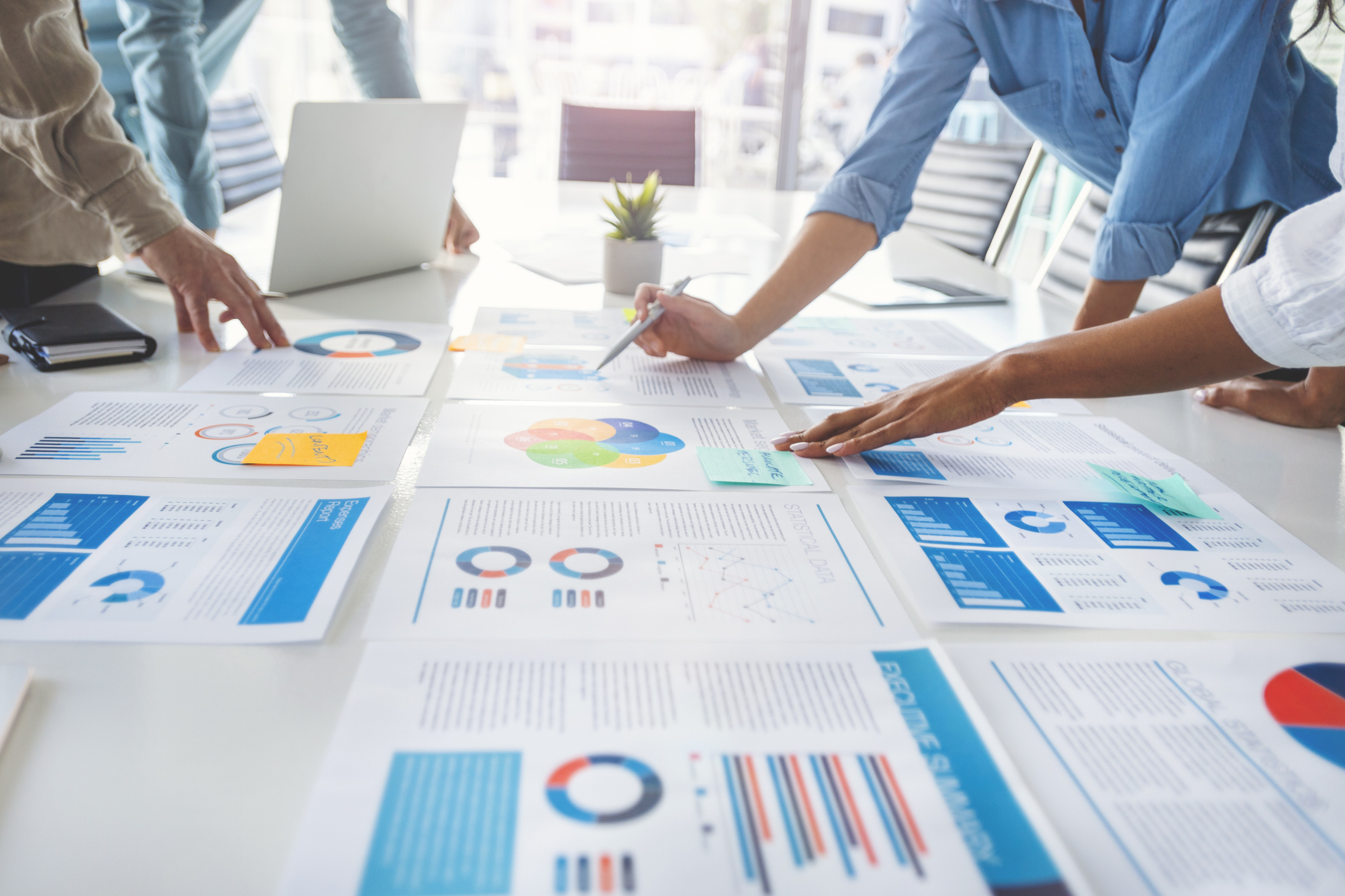 Paperwork and hands on a board room table at a business presentation or seminar. The documents have financial or marketing figures, graphs and charts on them. People are pointing to different documents. There is a laptop on the table