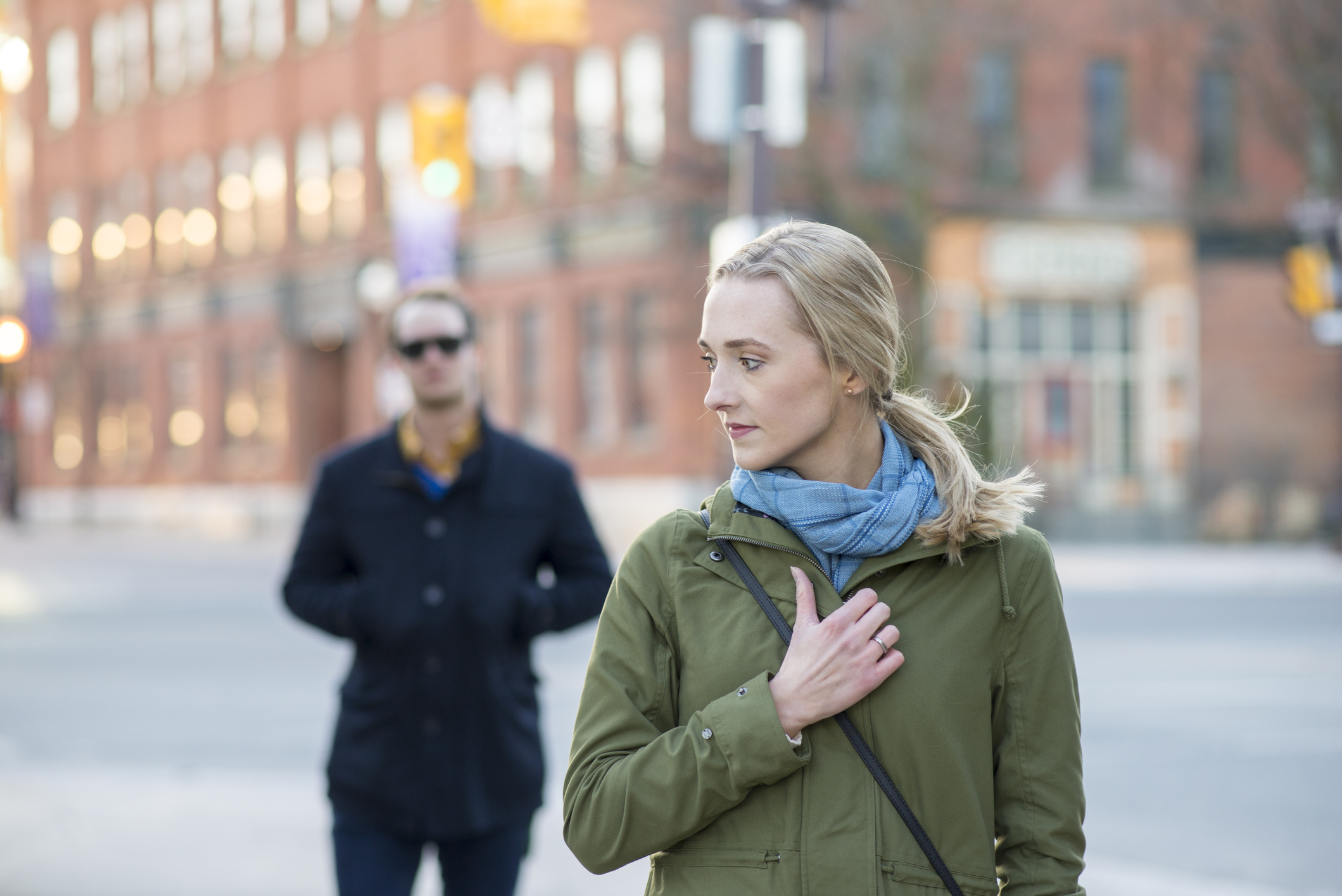 Scared young woman with blonde hair looking back over her shoulder at a stranger in a black trench coat that is following behind her downtown in an urban city.